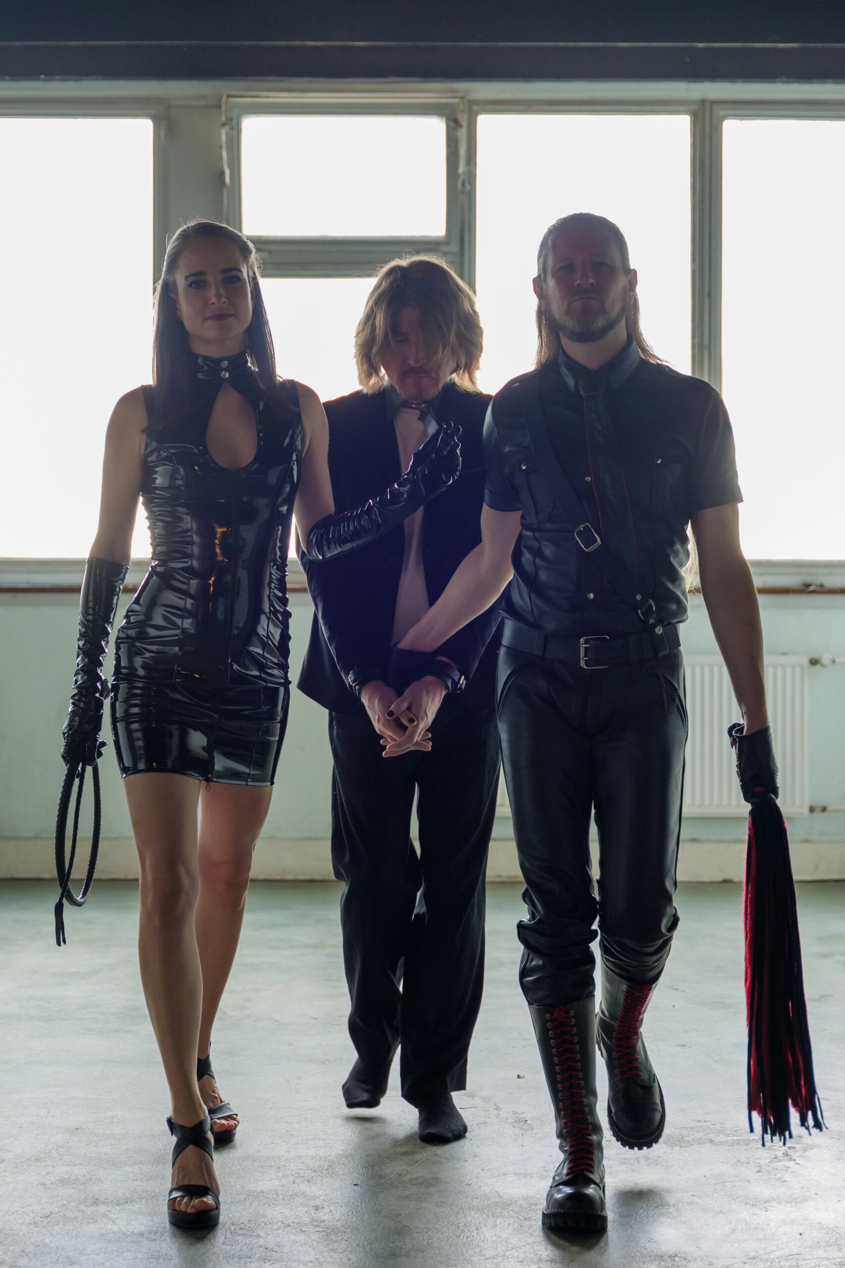 Hekate und Master Samael 11 – Leine Hekate in glänzendem Outfit mit Flogger links, am Halsband folgt ein männlicher Sub in schwarz. Recht hält Master Samael, in Leder, die Handfesseln des Subs und einen Flogger.