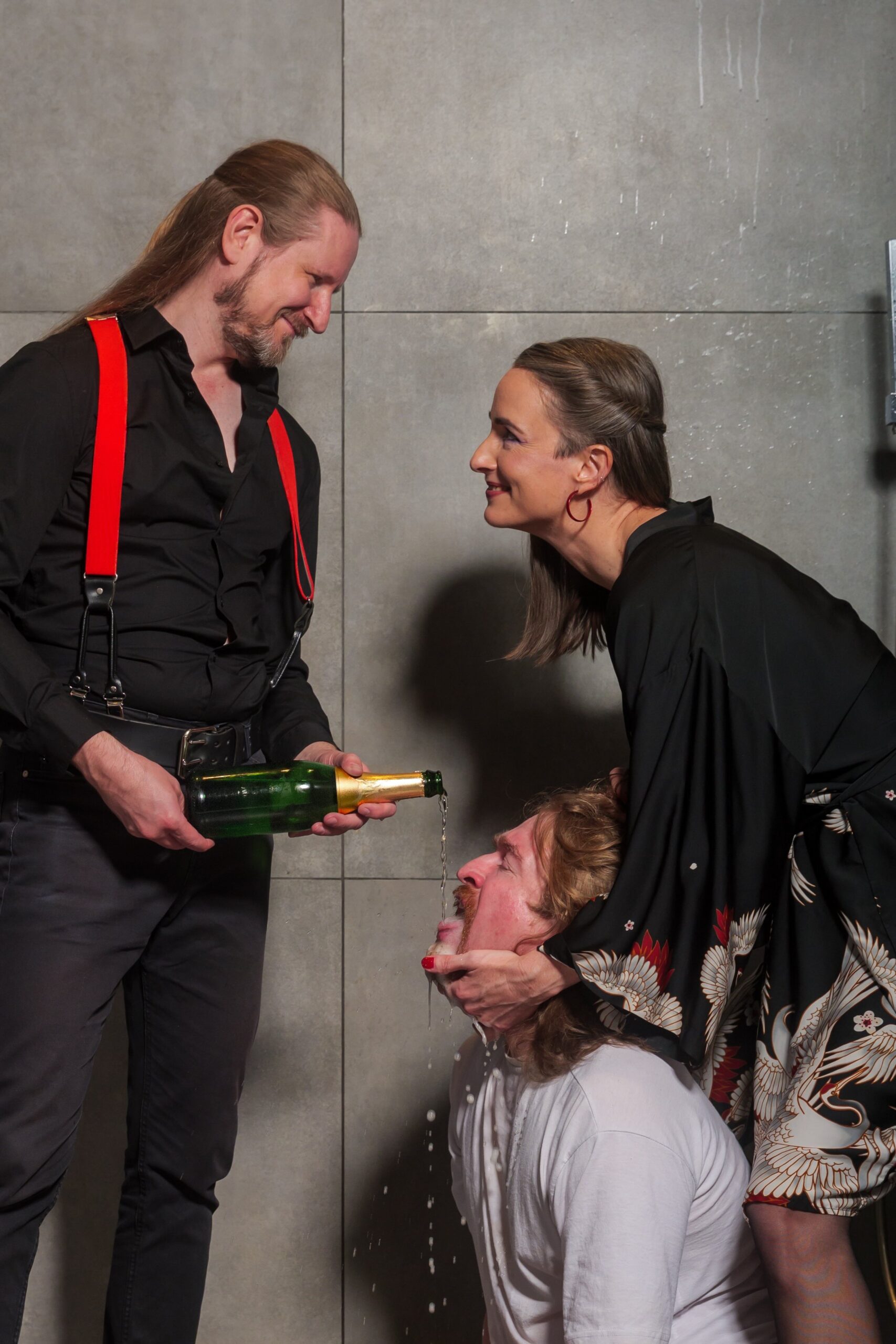 Hekate und Master Samael 9 – Lächelnd Master Samael im Hemd und Hekate im Negligé. Samael gießt eine Flasche frisch geköpften Champagner in das Gesicht eines knienden Subs. Hekate hält seinen Kopf und grinst.