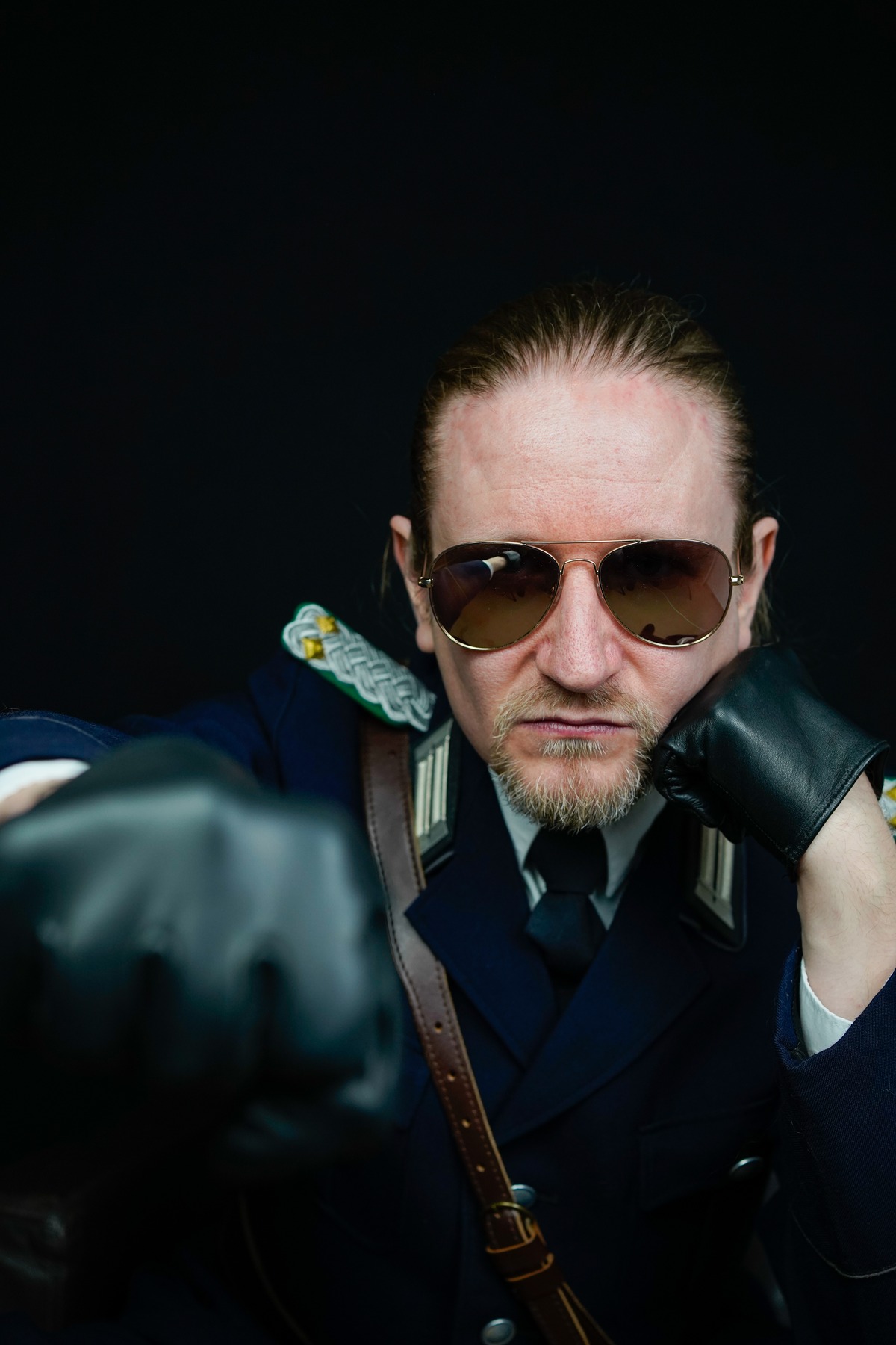 02_Master-Samael_Bild-2 Porträt-Foto von Master Samael, einem blonden Profi-Dom mit Sonnenbrille und schwarzen Lederhandschuhen. Eine Uniform ist auch erkennbar.