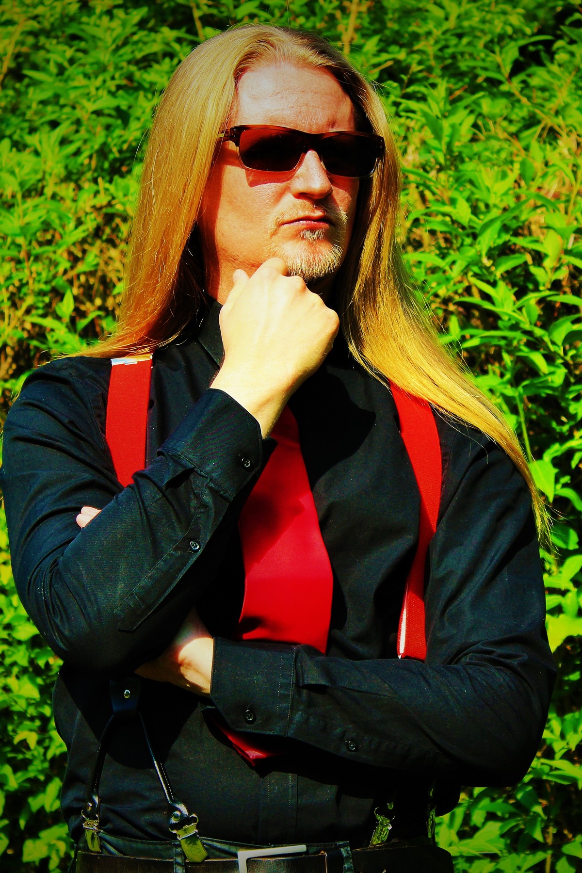 06_Master-Samael_Bild-6 Porträtfoto von Master Samael: Seine langen blonden Haare offen, mit Sonnenbrille, schwarzem Hemd sowie roter Krawatte und roten Hosenträgern vor einem grünen Busch