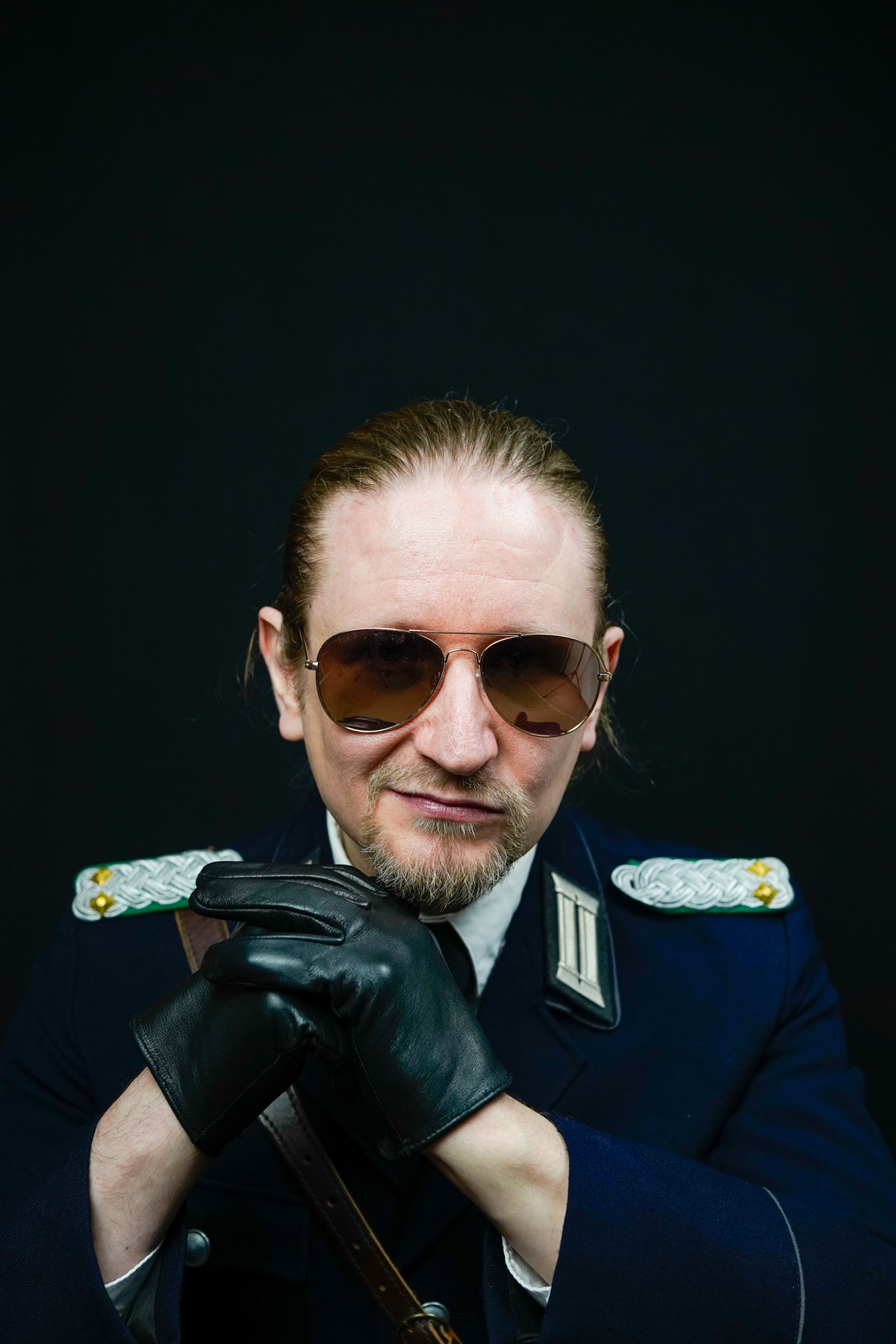 08_Master-Samael_Bild-8 Porträtfoto von Master Samael in einer Uniform mit schwarzen Lederhandschuhen und Sonnenbrille. Er grinst bedrohlich in die Kamera.